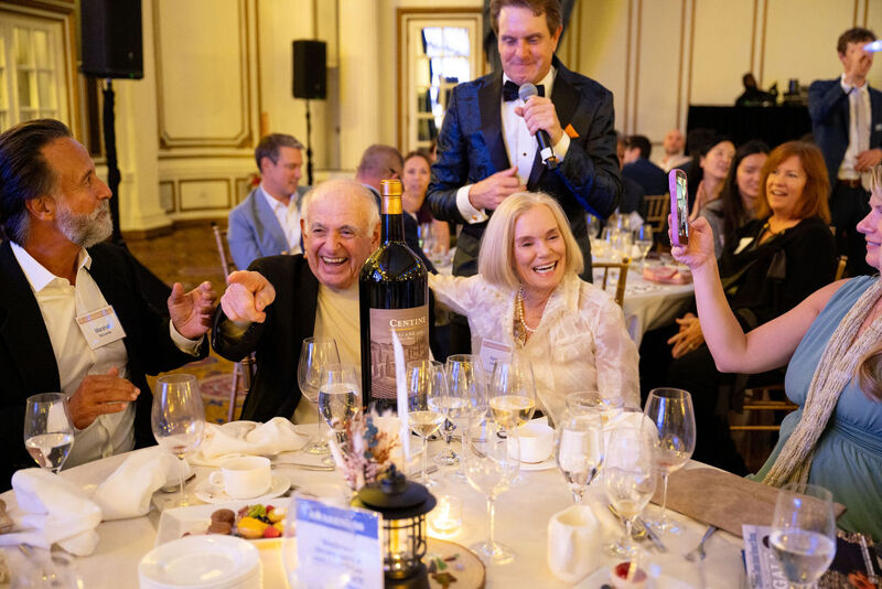 The image captures a lively dinner event. People are seated around a table, smiling and interacting. A man in a tuxedo is speaking into a microphone, possibly hosting or making an announcement. There's a bottle of wine on the table, suggesting a celebratory atmosphere. The overall mood is joyful and social.